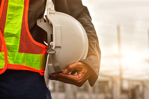 A person holding a white construction hat while wearing an orange-and-yellow safety vest over a grey long-sleeved shirt, and dark pants. A grey-and-white background with indistinct structures is blurred behind the individual.