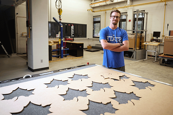 A picture of a white male student with his arms crossed, standing behind a large piece of cardboard on a worktable that has Sycamore leaf shapes cut out of it. The man has dark brown hair, a beard, and a moustache, and wears glasses. He is wearing a blue t-shirt that says STATE and is standing in an industrial lab. The top of the worktable is gray and is visible in the leaf cutouts.